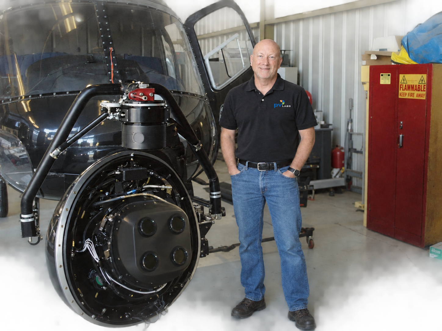 Ty Shattuck standing beside a PV Labs ISR gimbal sensor system mounted on a helicopter in a hangar